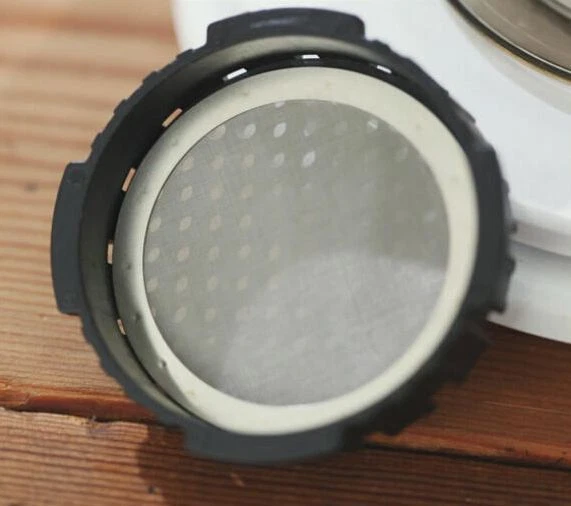 Stainless steel mesh AeroPress coffee filter sitting in filter basket on wooden surface, leaning against plates – reusable, eco-friendly
