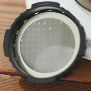 Stainless steel mesh AeroPress coffee filter sitting in filter basket on wooden surface, leaning against plates – reusable, eco-friendly